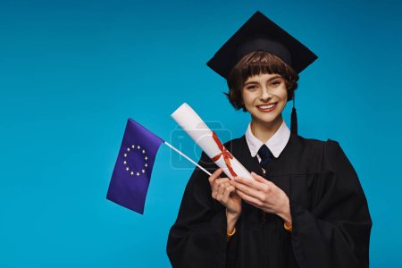 fröhliche Hochschulabsolventin in Kleid und akademischer Mütze mit Diplom und EU-Flagge auf blauem Hintergrund