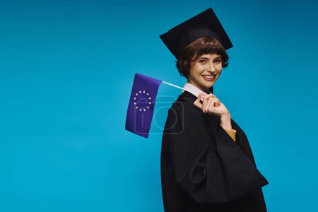 Hochschulabsolventin in Kleid und akademischer Mütze mit Diplom und EU-Flagge vor blauem Hintergrund lächelnd