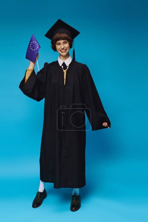 graduada chica universitaria en vestido y gorra académica sosteniendo la bandera de la UE y sonriendo en telón de fondo azul