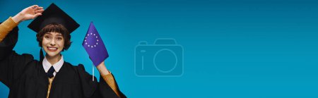 Photo for Graduated college girl in gown and academic cap holding EU flag and smiling on blue backdrop, banner - Royalty Free Image