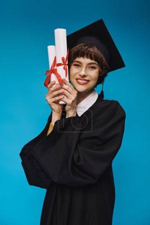 Eager grad college girl in gown and academic cap holding diplomas with pride, blue background