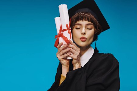 Téléchargez les photos : Heureux diplômé collège fille en robe et casquette académique détenant des diplômes avec fierté, fond bleu - en image libre de droit