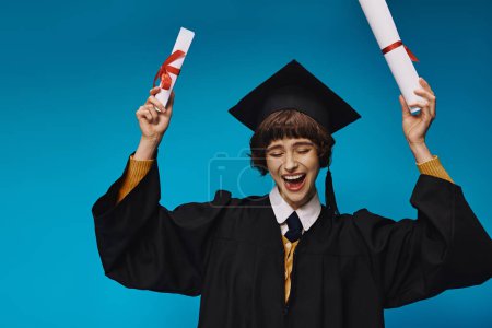 Photo for Eager grad college girl in gown and academic cap holding diplomas with pride, blue background - Royalty Free Image