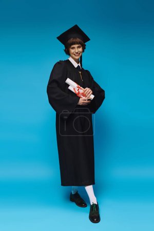 Photo for Happy grad college girl in gown and academic cap holding diplomas with pride, blue background - Royalty Free Image