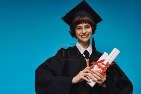 Foto de Feliz graduado chica universitaria en vestido y la gorra académica que sostiene diplomas con orgullo en azul - Imagen libre de derechos