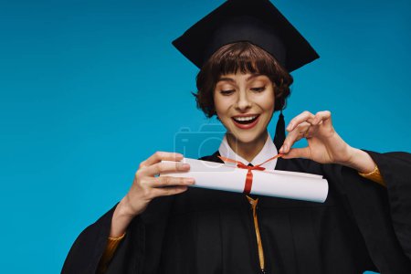 happy grad college girl in gown and academic cap looking at her diploma with pride on blue
