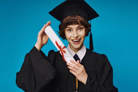 fröhliches College-Mädchen in Kleid und akademischer Mütze, das stolz sein Diplom auf blauem Grund hält, Abschluss