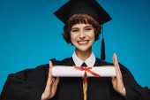 positive grad college girl in gown and academic cap holding her diploma with pride on blue Poster #712417856
