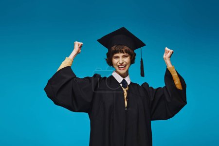 excited graduated college girl in gown and cap raising fists on blue background, accomplishment