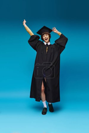 excited graduated college girl in gown and cap raising fists on blue background, success