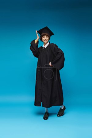 Photo for Excited graduated college girl in gown and cap smiling on blue background, accomplishment - Royalty Free Image