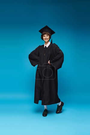 Photo for Cheerful graduated college girl in gown and cap smiling on blue background, accomplishment - Royalty Free Image