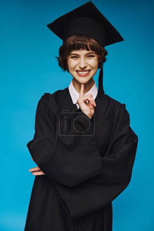 Foto de Chica universitaria graduada positiva en vestido y gorra sonriendo sobre fondo azul, logro - Imagen libre de derechos