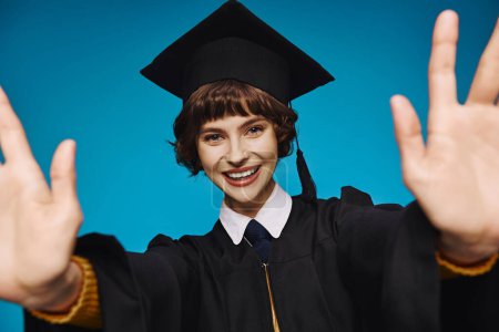 Photo for Happy graduate college girl in academic cap with outstretched hands on blue backdrop, accomplishment - Royalty Free Image
