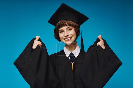 cheerful graduate college girl in academic cap making heart sign with her fingers on blue backdrop Poster 712418118