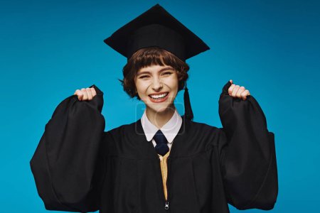 Photo for Young brunette graduate college girl in academic cap feeling happy on blue background, success - Royalty Free Image
