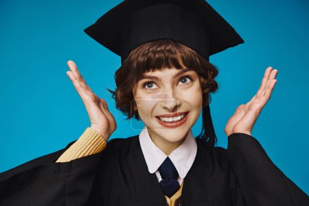 Photo for Young and cheerful graduate college girl in academic cap gesturing with hands, blue background - Royalty Free Image