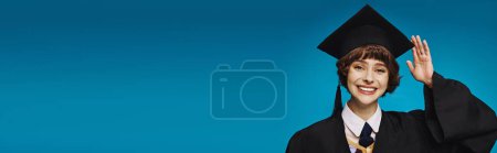 Photo for Young and cheerful graduate college girl in academic cap saluting with hand, blue background banner - Royalty Free Image