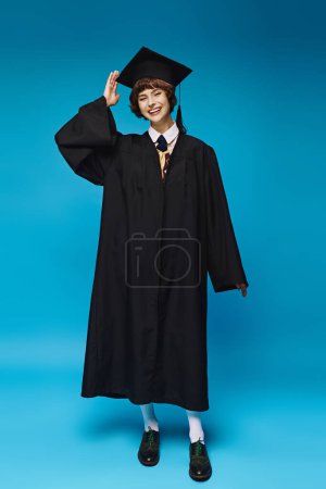Photo for Excited young woman in black graduation gown and academic cap smiling on blue background, ceremony - Royalty Free Image