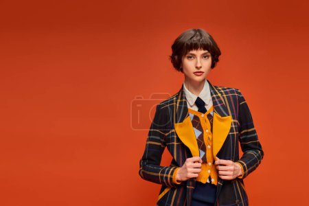 stylische Studentin mit kurzen Haaren posiert im karierten Blazer auf orangefarbenem Hintergrund, College-Uniform