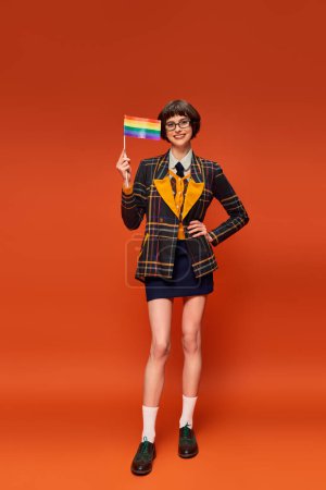 Foto de Feliz joven universitaria en uniforme y gafas con bandera lgbt y de pie sobre fondo naranja - Imagen libre de derechos