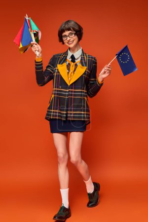 happy young college girl in uniform and glasses holding EU and different flags on orange background