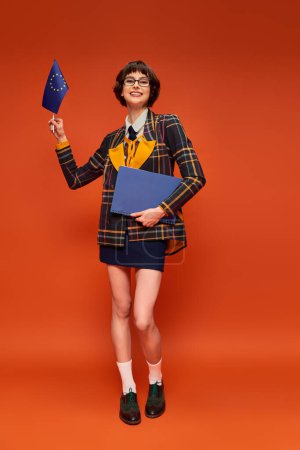 heureuse jeune fille de l'université en uniforme et lunettes tenant drapeau de l'UE avec ordinateur portable sur fond orange