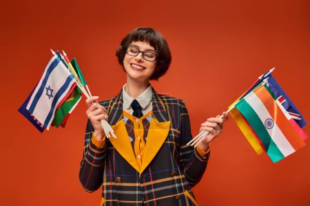 chica universitaria feliz en su uniforme y gafas con múltiples banderas y de pie sobre fondo naranja
