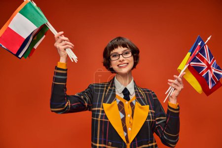 Téléchargez les photos : Heureuse étudiante fille dans son uniforme et lunettes tenant plusieurs drapeaux et debout sur fond orange - en image libre de droit