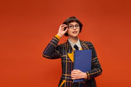 Estudiante reflexiva en uniforme universitario ajustando sus gafas y sosteniendo el cuaderno sobre fondo naranja