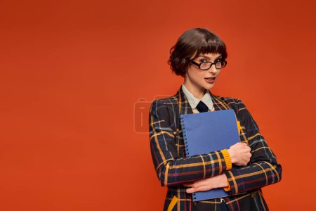 Thoughtful student in college uniform and glasses holding notebook on orange background, smart girl