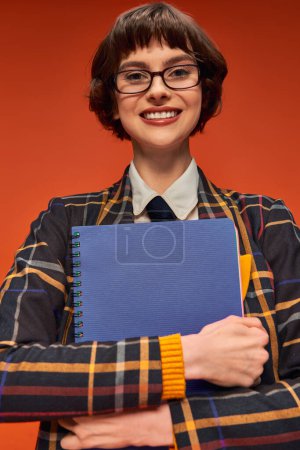 happy student in checkered uniform and glasses holding notebook on orange background, college girl