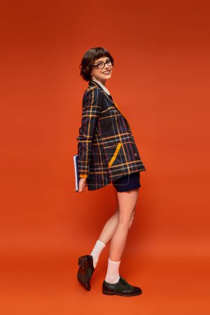 glücklicher Student in College-Uniform und Brille mit Notizbuch auf orangefarbenem Hintergrund, intelligentes Mädchen