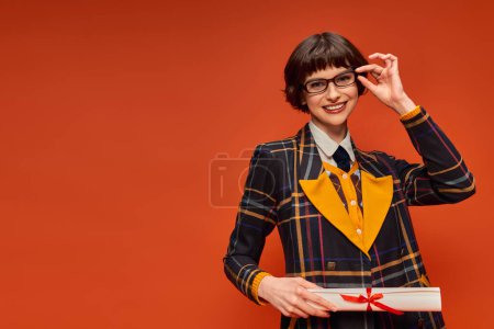 fröhliches Hochschulmädchen in Uniform und Brille mit Diplom vor leuchtend orangefarbenem Hintergrund