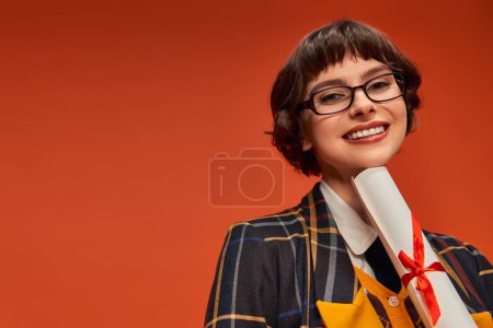 chica universitaria positiva en uniforme y gafas que sostienen su diploma de graduación en fondo naranja