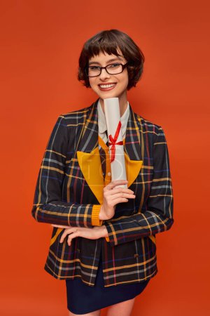 smiling college girl in uniform and glasses holding her graduation diploma on orange backdrop