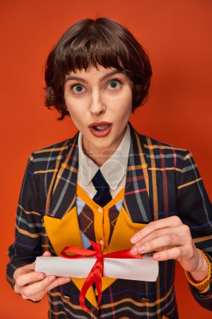 portrait of shocked college girl in checkered uniform holding graduation diploma on orange backdrop