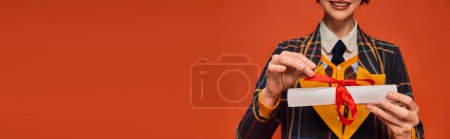 Photo for Cropped banner of happy college girl in checkered uniform holding graduation diploma on orange - Royalty Free Image