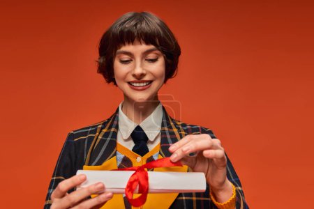 Foto de Retrato de chica universitaria feliz en uniforme a cuadros mirando diploma de graduación en fondo naranja - Imagen libre de derechos