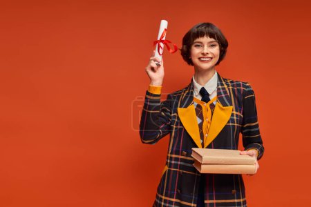 portrait of cheerful student in college uniform holding books and diploma on orange backdrop tote bag #712419992
