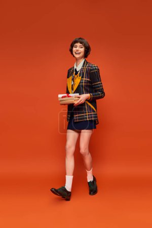 portrait of positive student in college uniform holding books and diploma on orange backdrop Stickers 712420010