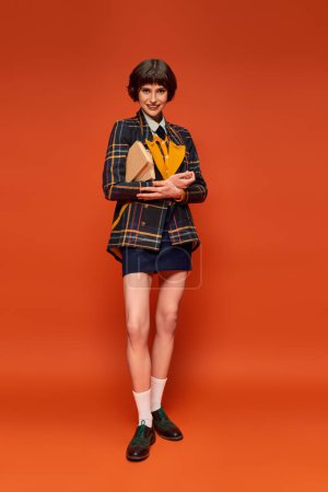 portrait of positive student in college uniform standing with books on orange background, knowledge