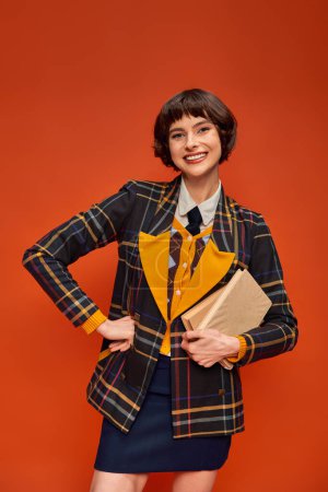 Porträt eines lächelnden Studenten in College-Uniform mit Büchern auf orangefarbenem Hintergrund, Wissen