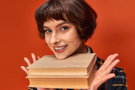 portrait of smiling college girl in uniform holding books near chin on orange background, knowledge mug #712420084