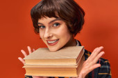 portrait of smiling college girl in uniform holding books near chin on orange background, knowledge t-shirt #712420084