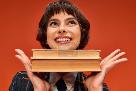 Porträt eines fröhlichen College-Mädchens in Uniform mit Büchern am Kinn auf orangefarbenem Hintergrund, Wissen