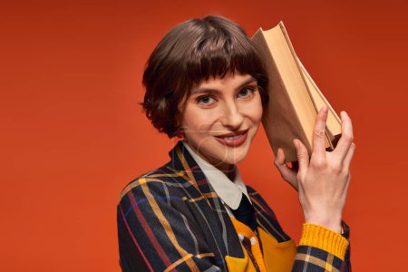 portrait of cheerful college girl in uniform holding books near face on orange background, knowledge