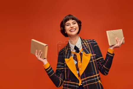 portrait of cheerful college girl in uniform holding books in hands on orange background, knowledge