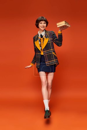 cheerful college girl in uniform holding stack of books in hand on orange background, knowledge mug #712420232