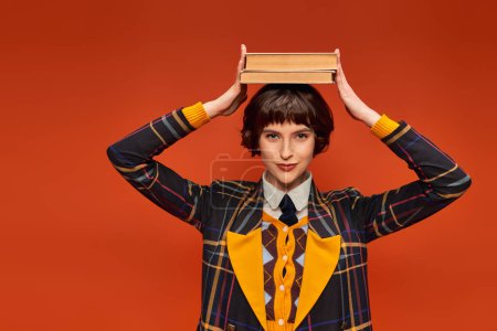 happy college girl in uniform holding stack of books on hand on orange background, knowledge